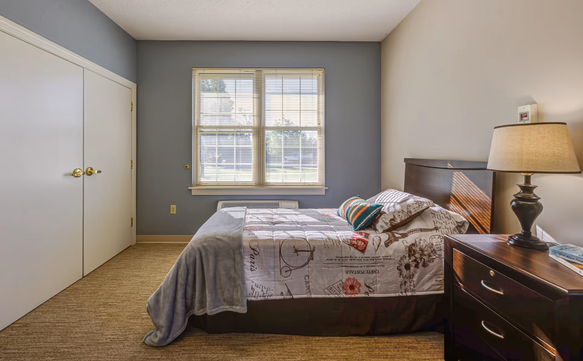 A bedroom with a bed against the right wall, covered with a patterned bedspread and a colorful pillow. Next to the bed is a dark wooden nightstand with a lamp and some books. A window with blinds is centered on the back wall, letting in natural light. On the left side, there is a closed double-door closet. The walls are painted in two tones, gray and beige, and the floor is carpeted.