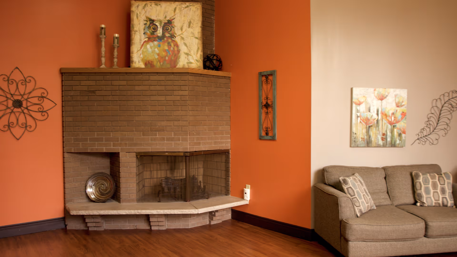 Cozy living room with a brick fireplace, orange accent wall, and a gray sofa with patterned pillows.