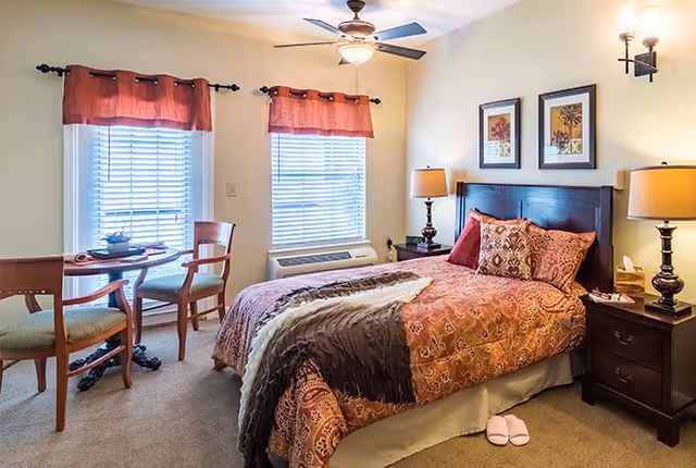 A cozy bedroom with a double bed covered in a patterned orange and brown bedspread, two matching pillows, and a gray throw blanket. There are two wooden nightstands with lamps on either side of the bed. Two windows with red valances let in natural light. A small round table with two chairs is positioned near the windows. Pink slippers are placed on the carpeted floor next to the bed.