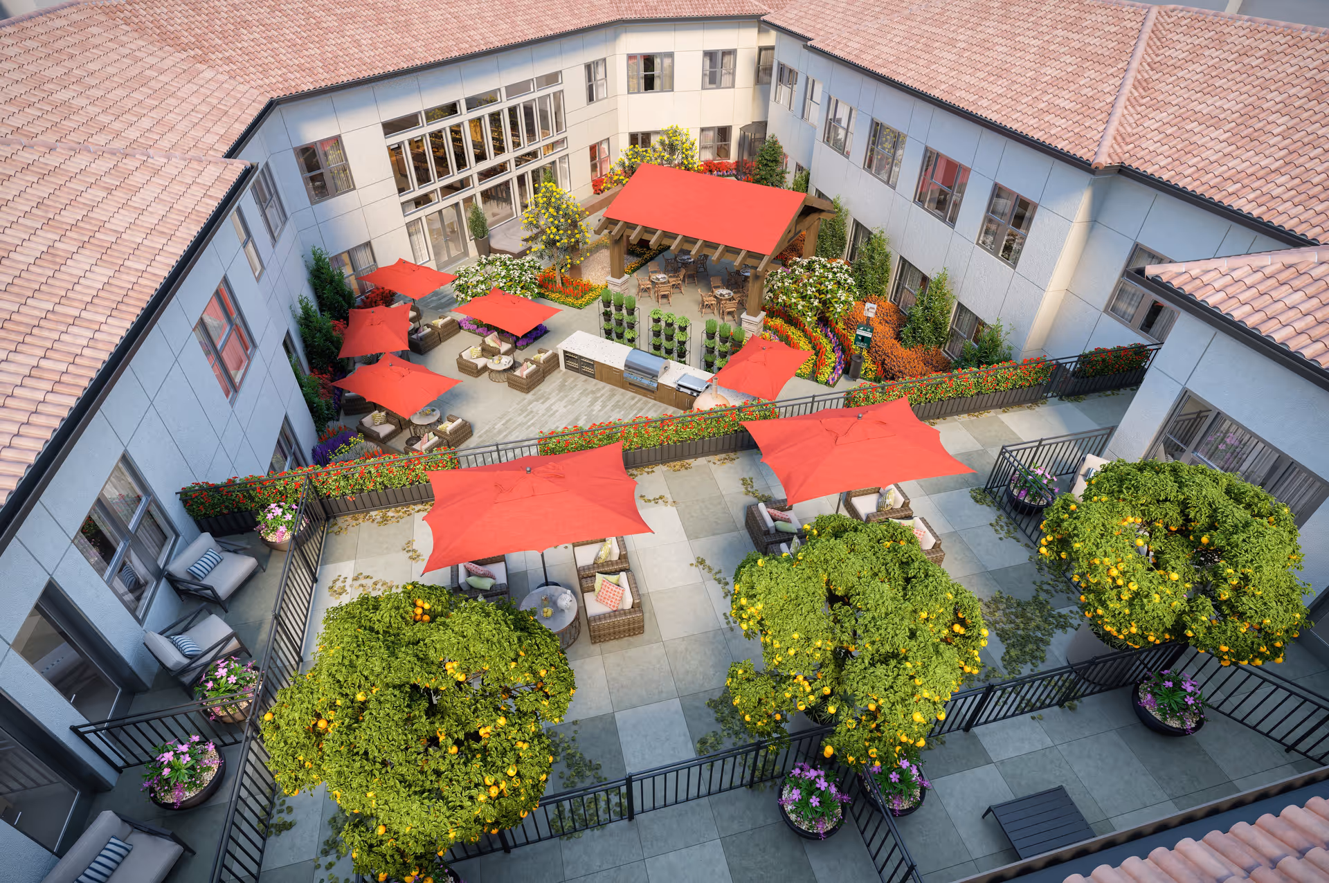 Aerial view of an outdoor courtyard at Varenita Simi Valley featuring multiple seating areas with red umbrellas, lush greenery including orange trees, colorful flower beds, and a covered pavilion with tables and chairs. The courtyard is surrounded by a building with tiled roofs and large windows.
