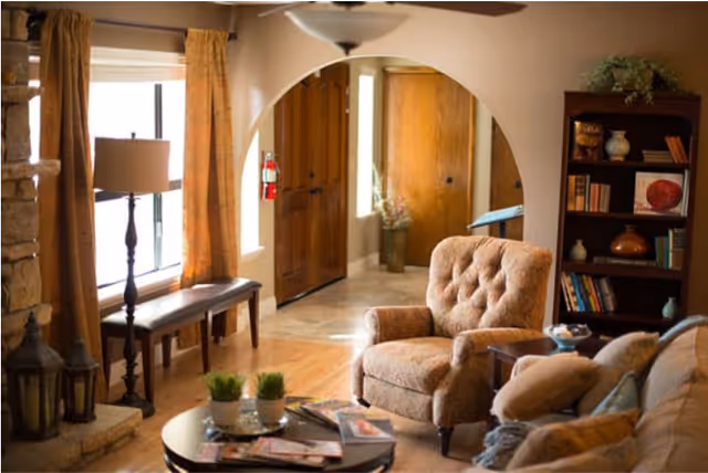 Cozy living room with an upholstered armchair and sofa around a coffee table, bookshelf, arched doorway, and window with curtains.