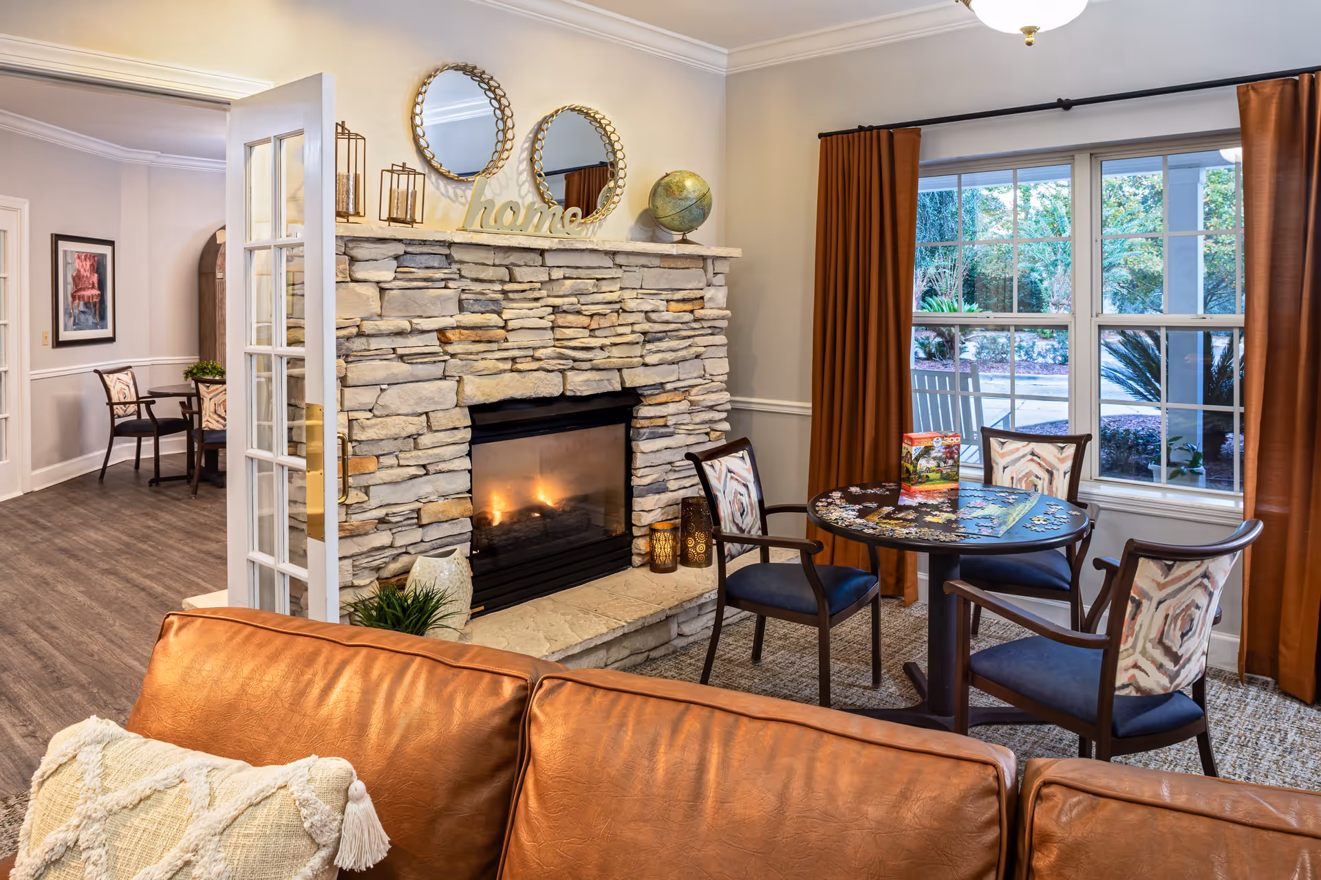Cozy living room area with a stone fireplace lit with a warm fire, decorated with two round mirrors, a globe, and the word 'home' on the mantel. In front of the fireplace is a round table with a puzzle in progress and four chairs with patterned backs. Large windows with brown curtains show a view of greenery outside. A brown leather sofa with a textured cream pillow is partially visible in the foreground.