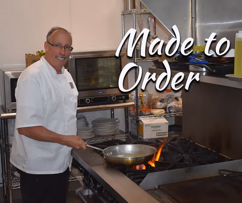 A chef in a white uniform cooking with a frying pan over a gas stove in a kitchen. Behind him are shelves with plates, kitchen equipment, and food supplies. The words 'Made to Order' are overlaid on the right side of the image.