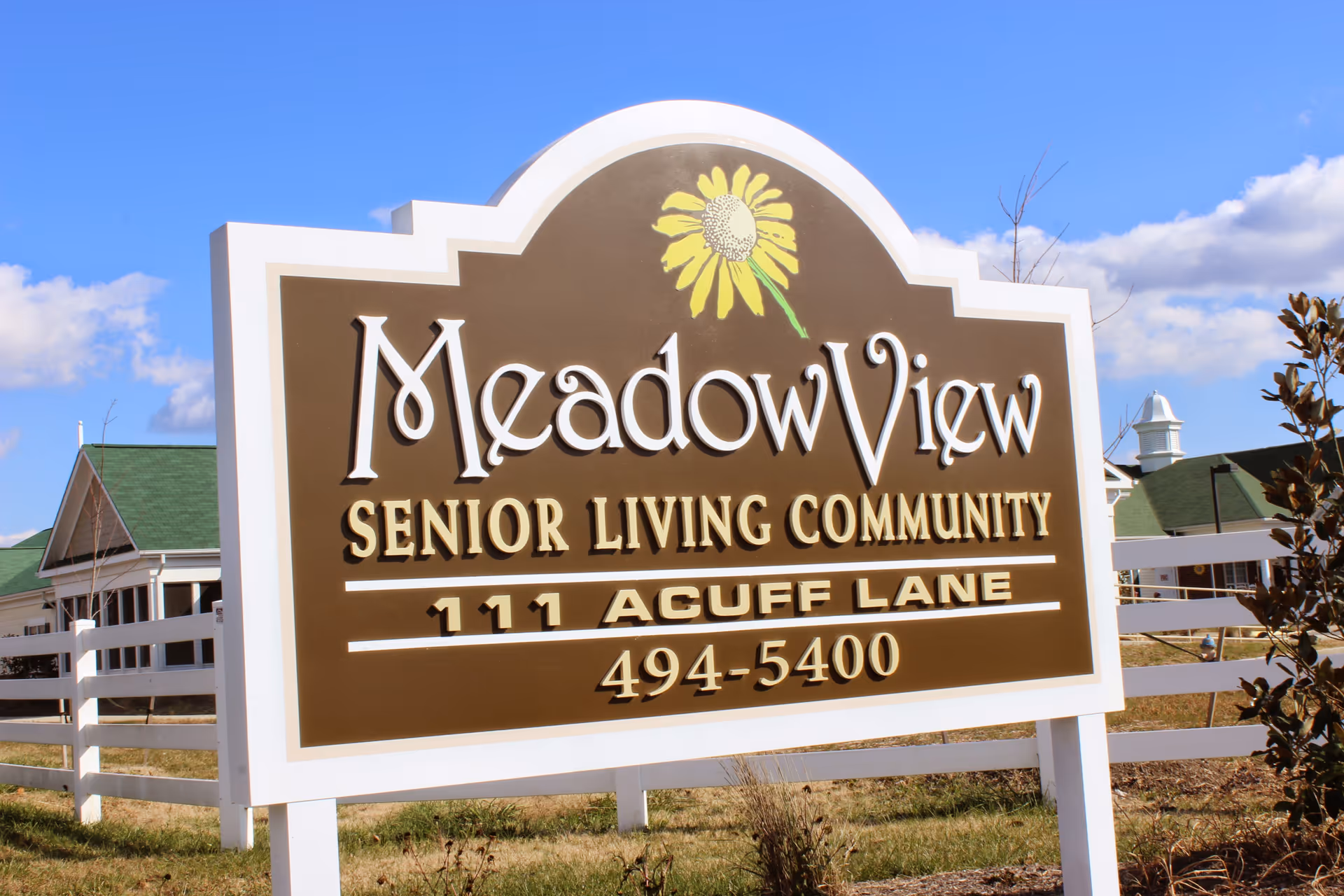 A large outdoor sign for Meadow View Senior Living Community with a yellow flower graphic, located at 111 Acuff Lane, with a phone number 494-5400. The background shows part of the facility buildings and a white fence under a blue sky with some clouds.