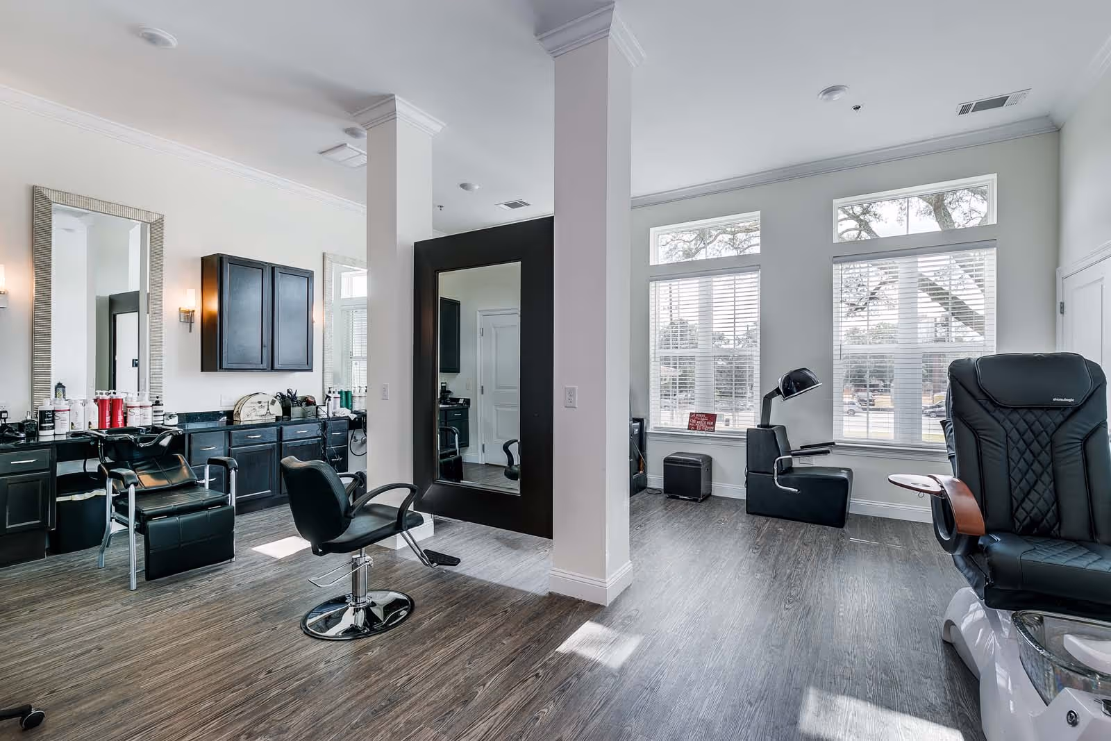 A modern salon area with black styling chairs, large mirrors, and black cabinetry along the wall. The room has large windows with white blinds allowing natural light to fill the space. There is a pedicure chair on the right side near the windows and a small seating area with a lamp and ottoman in the corner.