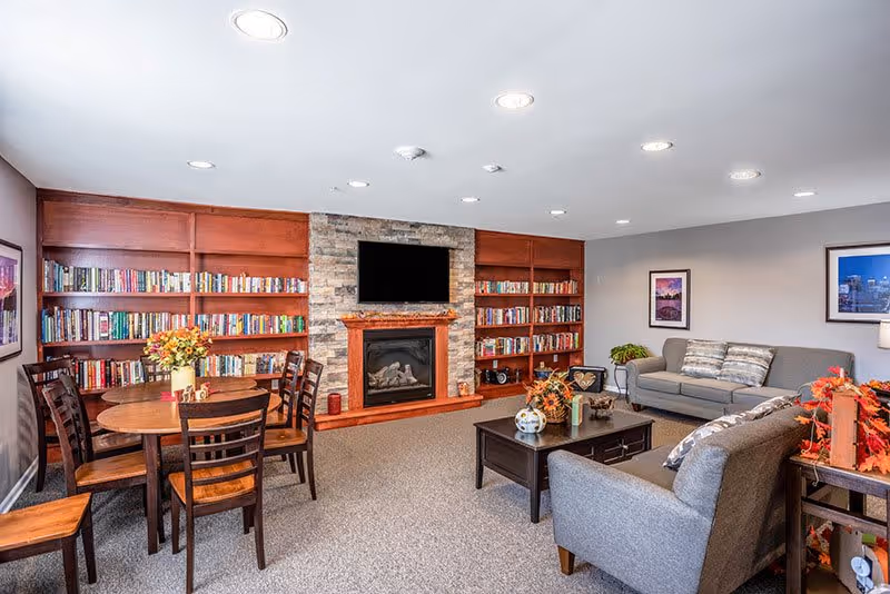 A cozy living room area with a stone fireplace and a mounted flat-screen TV above it. The fireplace is flanked by wooden bookshelves filled with books. There is a round wooden table with four chairs on the left side, decorated with a vase of flowers. Two gray sofas with cushions face a dark wooden coffee table adorned with decorative items. The walls are light-colored with framed pictures, and the room is well-lit with recessed ceiling lights.