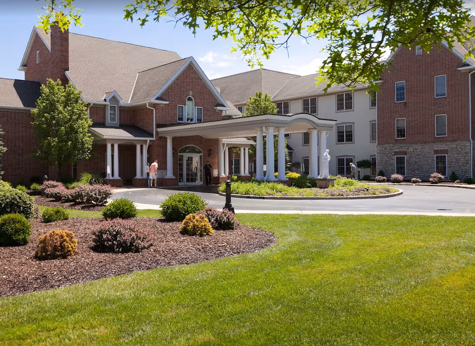 Front exterior view of Ancora at Mount Alverna Village, showing a large brick and siding building with a covered entrance supported by white columns. There are landscaped bushes and trees in front, with a green lawn and a circular driveway. A few people are near the entrance.