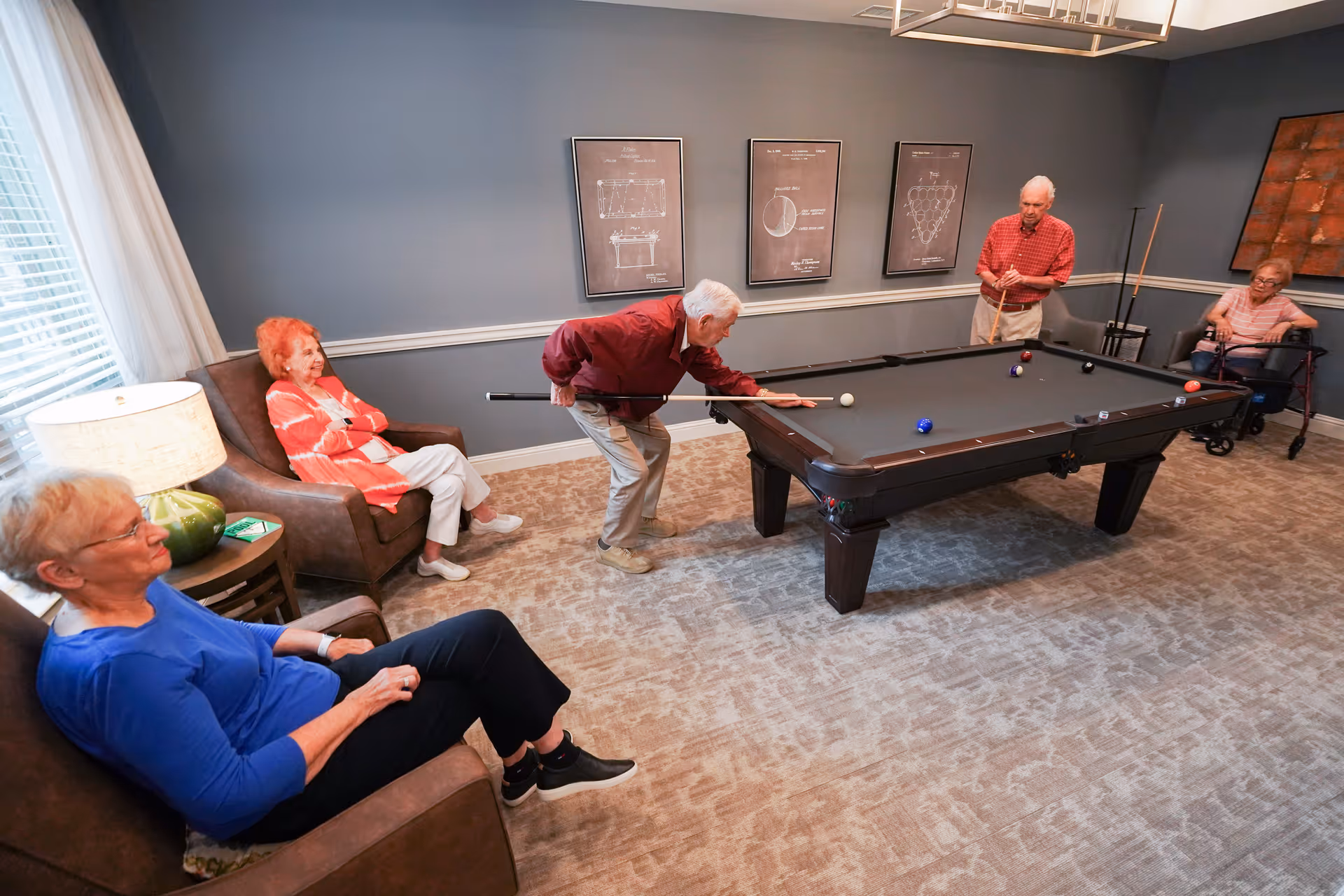 A group of elderly people in a common room playing and watching a game of pool. One man is taking a shot while another man stands at the opposite side of the pool table holding a cue stick. Two women are seated nearby, one in a brown armchair and the other in a wheelchair. The room has gray walls, carpeted floor, and framed artwork on the walls.