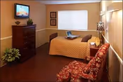A cozy bedroom in a nursing and rehabilitation center featuring a bed with a brown quilt, a wooden dresser with a TV on top, a red patterned armchair, a small nightstand with a lamp, and a window with closed blinds.