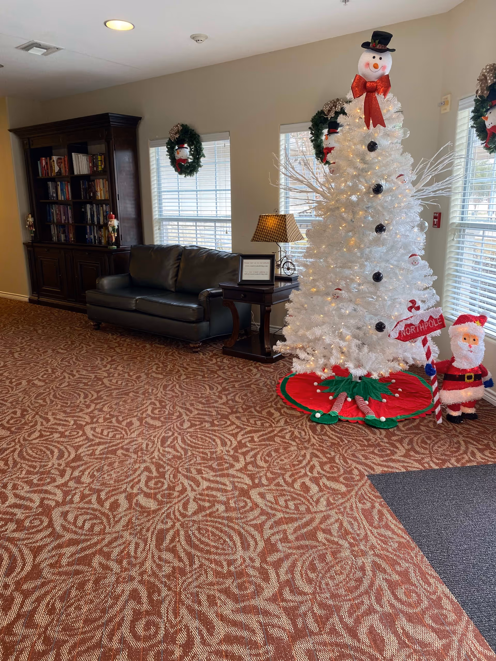Interior of a senior living facility decorated for Christmas with a white Christmas tree adorned with snowman ornaments and a red bow on top, a small Santa figure holding a North Pole sign, a dark leather couch, a wooden bookshelf filled with books, two windows with Christmas wreaths hanging, and a side table with a lamp and a framed sign.
