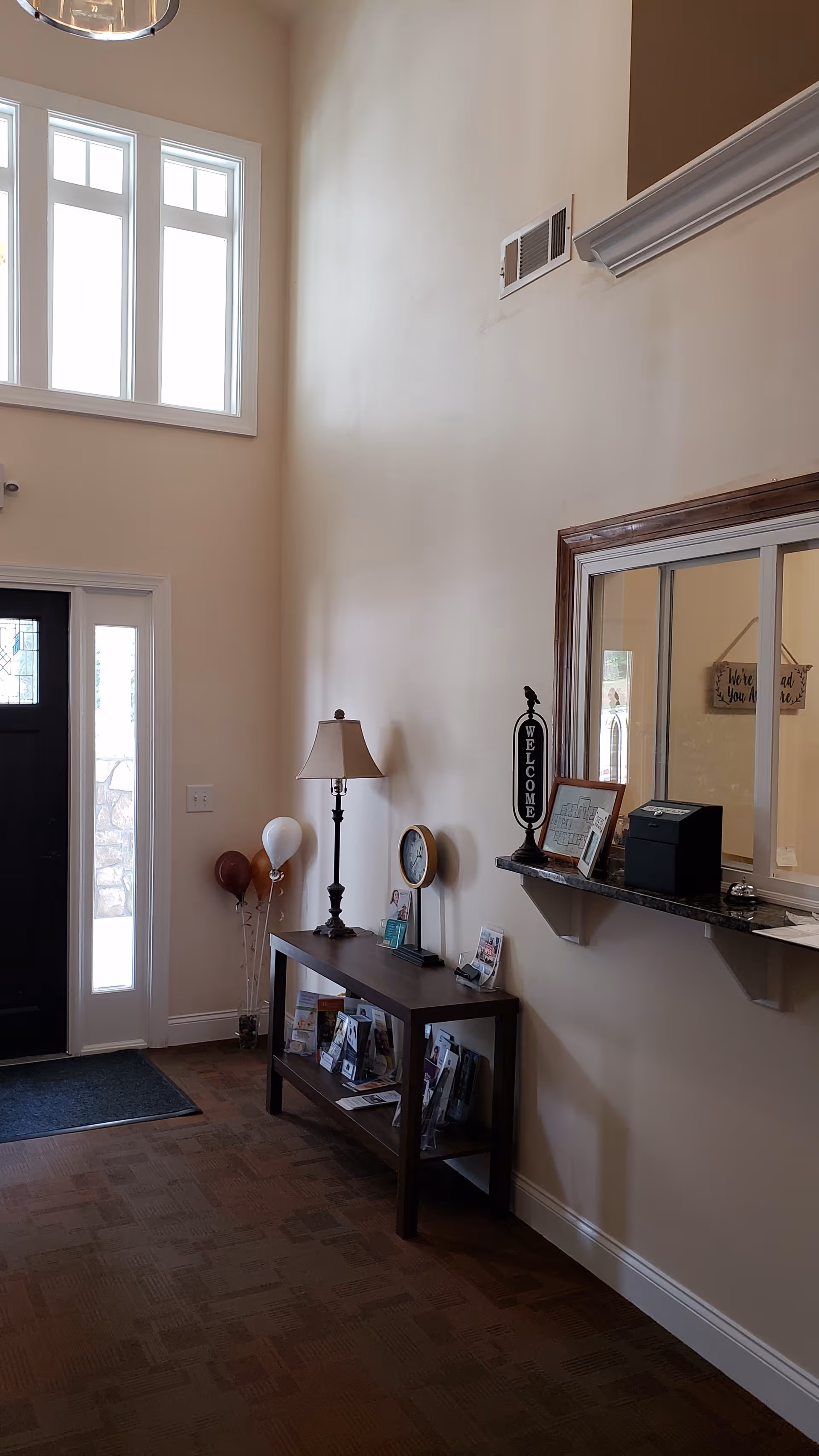 Bright two-story entry foyer with a front door, console table holding a lamp and brochures, and a reception window with a small counter.