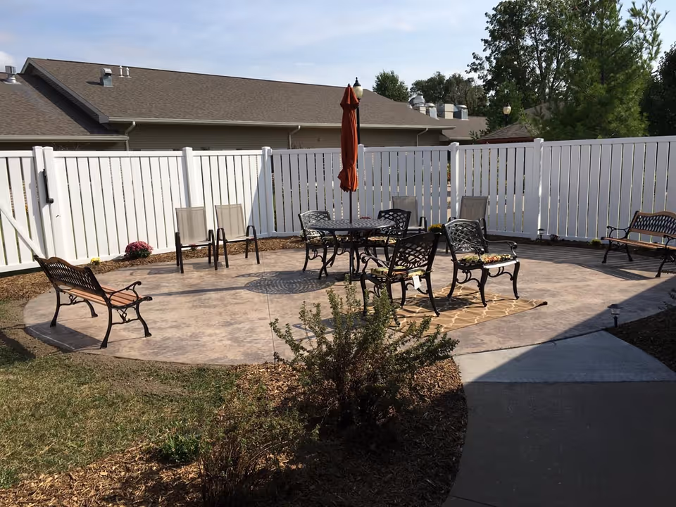 Outdoor patio area with a circular concrete seating space surrounded by a white privacy fence. The patio has several metal benches and chairs, a round table with an umbrella in the center, and some small plants and mulch around the perimeter.