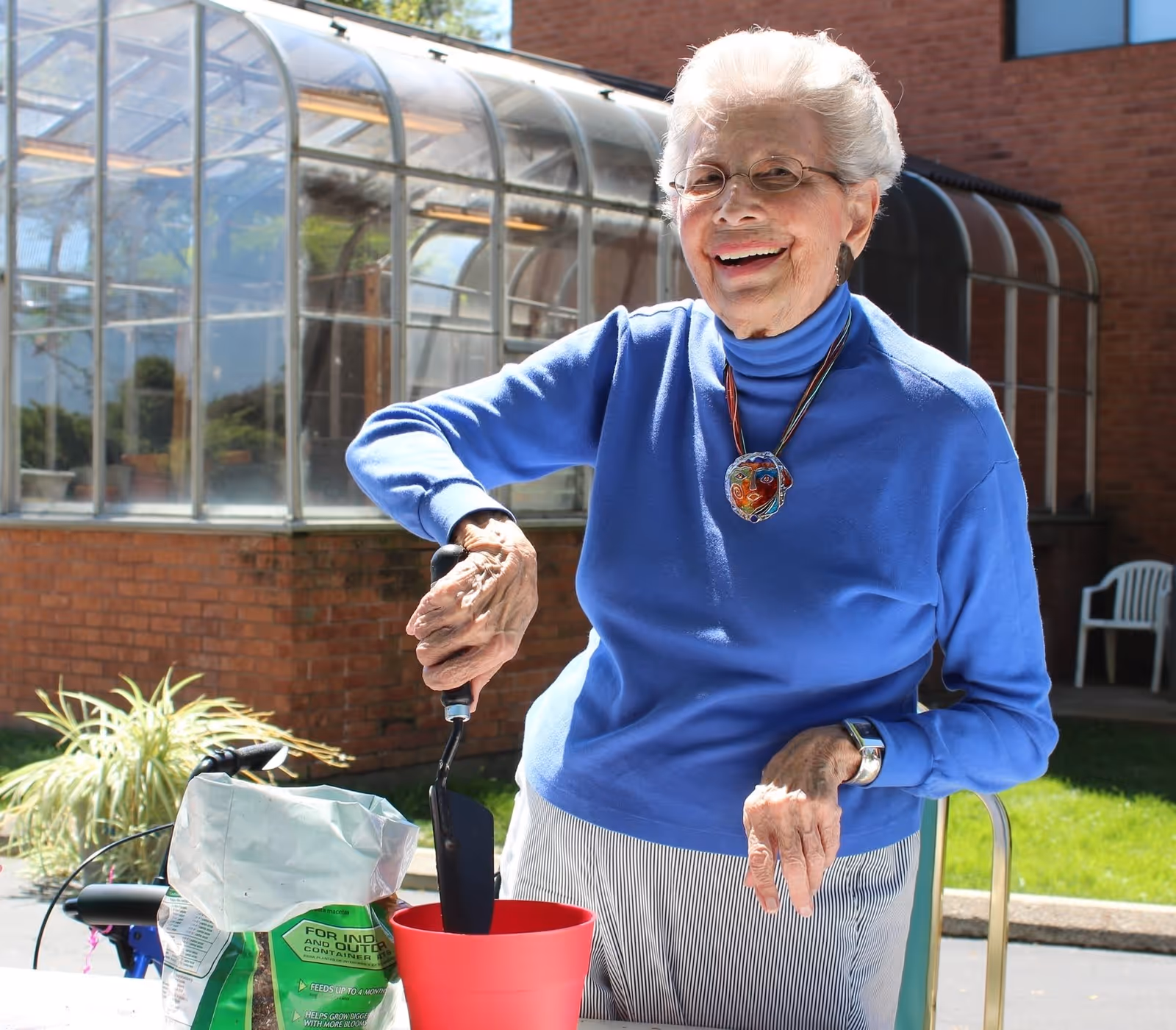 An elderly woman wearing glasses, a blue turtleneck sweater, and striped pants is smiling while gardening outdoors. She is holding a small gardening trowel and standing next to a red flower pot and a bag of potting soil. Behind her is a greenhouse structure and a brick building.