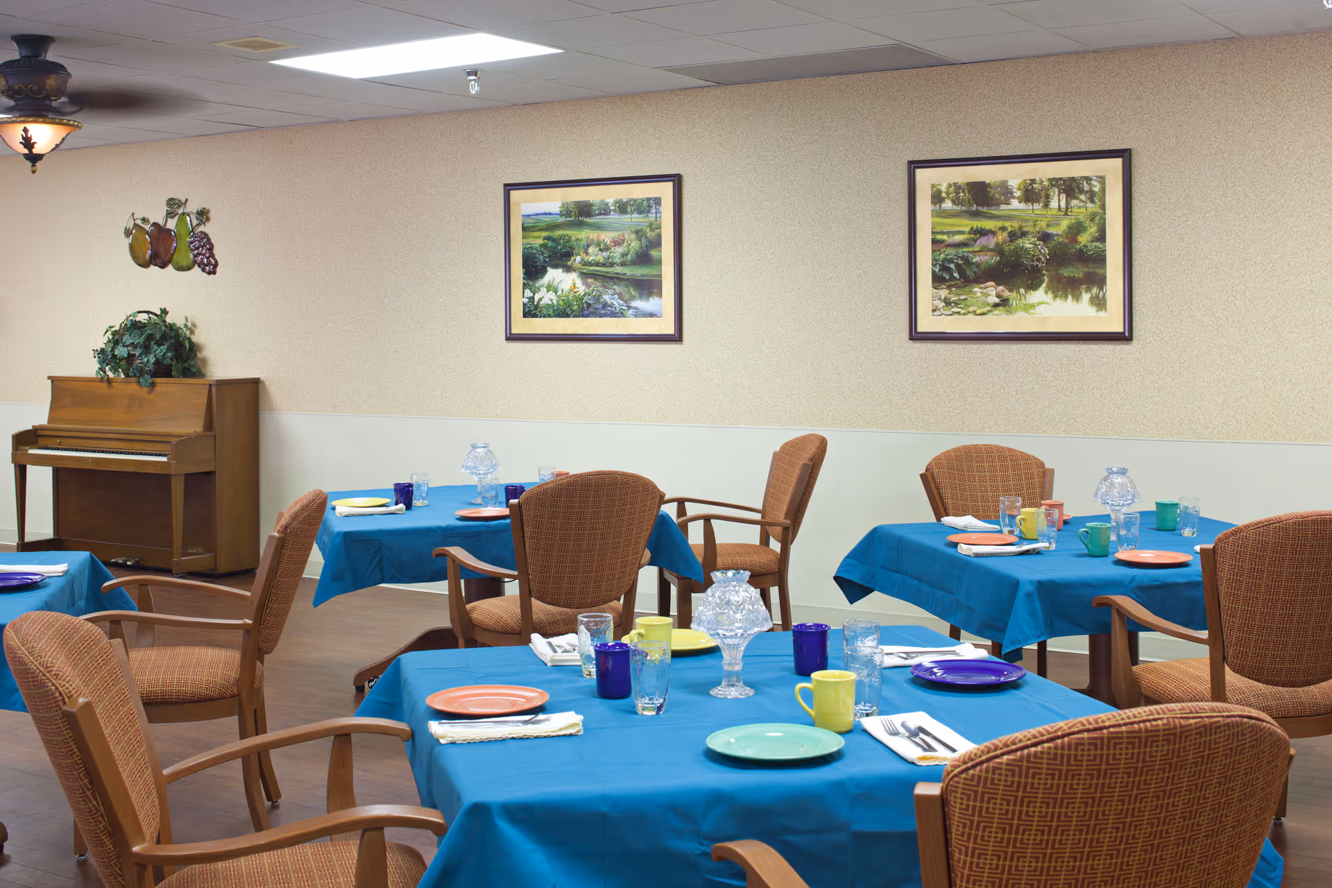 Dining room with tables covered in blue tablecloths set with colorful plates and glasses, surrounded by upholstered chairs and a piano against the wall.
