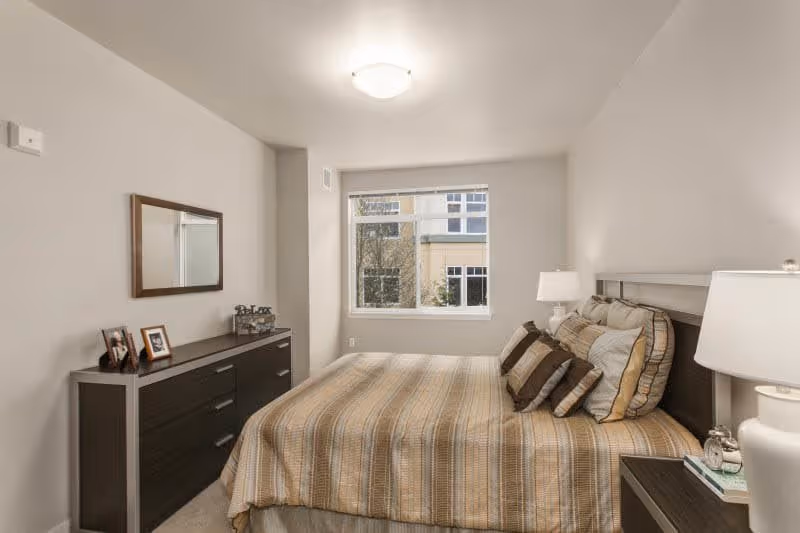 A neatly arranged bedroom with a large bed covered in a striped beige and brown bedspread, multiple pillows, a dark wooden headboard, and matching nightstands with lamps on either side. A window at the far end lets in natural light, and a dark wooden dresser with framed photos and decorative items is positioned against the left wall beneath a rectangular mirror.