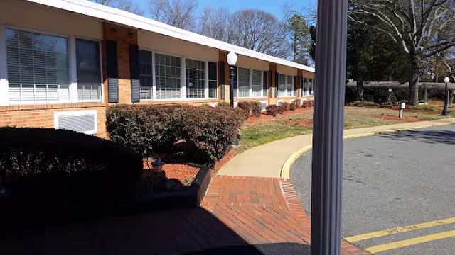 Exterior view of a single-story brick building with large windows and black shutters, surrounded by trimmed bushes and a curved sidewalk under a clear blue sky.