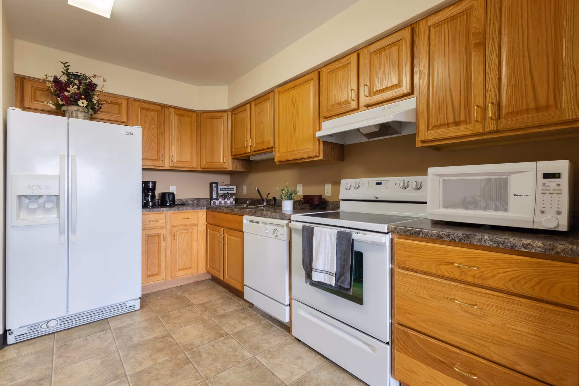 A kitchen with wooden cabinets, a white refrigerator with an ice and water dispenser, a white dishwasher, a white stove with a towel hanging on the handle, and a white microwave on the countertop. There are small appliances like a coffee maker and toaster on the counter, along with decorative items including a flower arrangement and a small potted plant.