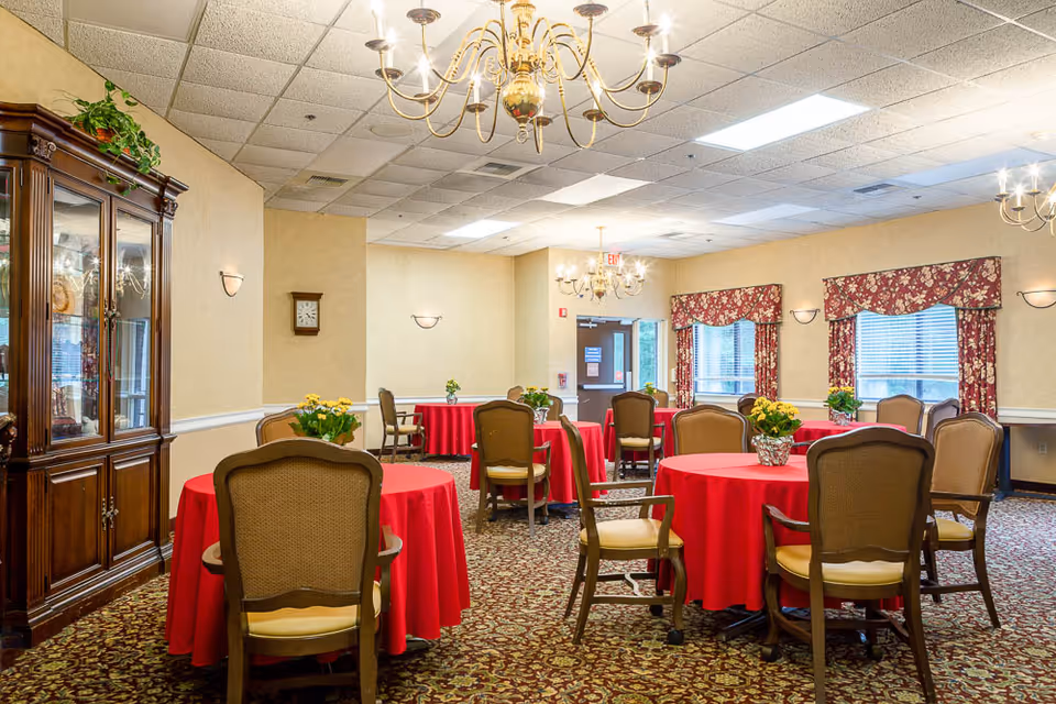 Dining room with round tables draped in red tablecloths, floral centerpieces, and upholstered chairs under chandeliers.