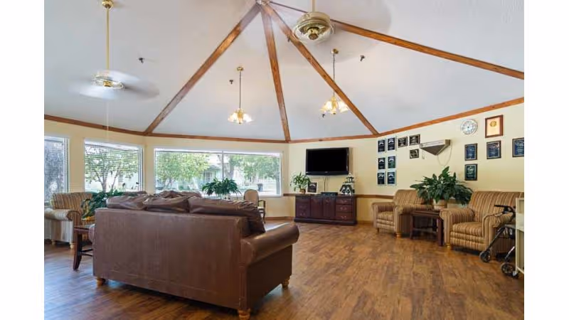 A spacious living room with a high ceiling featuring wooden beams and ceiling fans. The room has large windows letting in natural light and is furnished with a brown leather sofa, striped armchairs, wooden side tables, and a TV mounted on the wall above a wooden cabinet. Several plants and framed awards or certificates decorate the room.