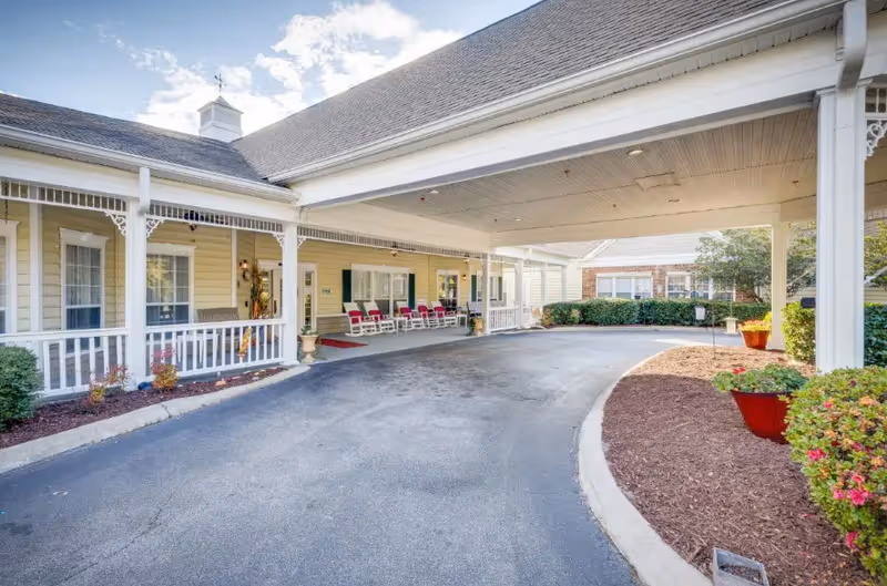 Covered driveway entrance to a senior living facility with a yellow exterior, white railings, and several red rocking chairs on the porch. There are landscaped areas with bushes and flowers around the driveway.