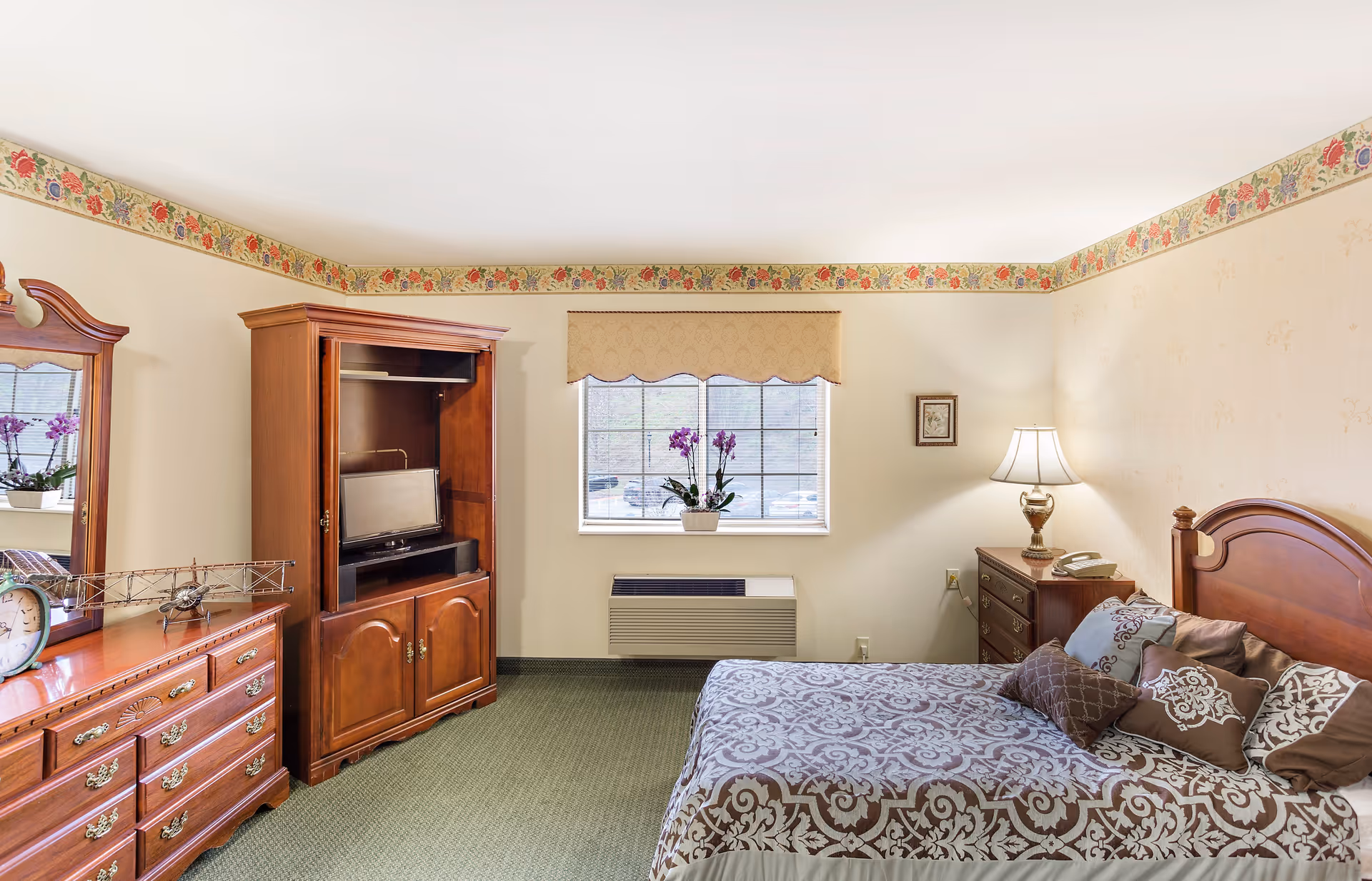 Bedroom with a wooden bed and matching furniture, patterned bedspread, dresser and armoire with a TV, and a window with a potted orchid.