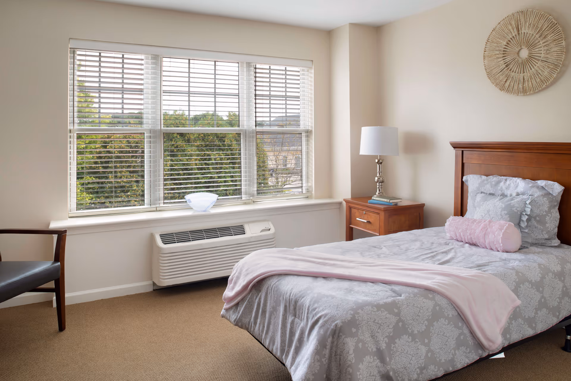 A bright bedroom with a single bed dressed in gray patterned bedding, a wooden nightstand and lamp, a chair, and a large window with blinds showing trees outside.