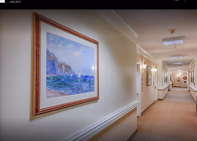A well-lit hallway in a senior living facility with beige walls and carpeted floor. The hallway features framed artwork on the walls, including a large painting of a coastal scene with cliffs and sailboats. There are handrails along the walls and ceiling lights evenly spaced along the corridor.