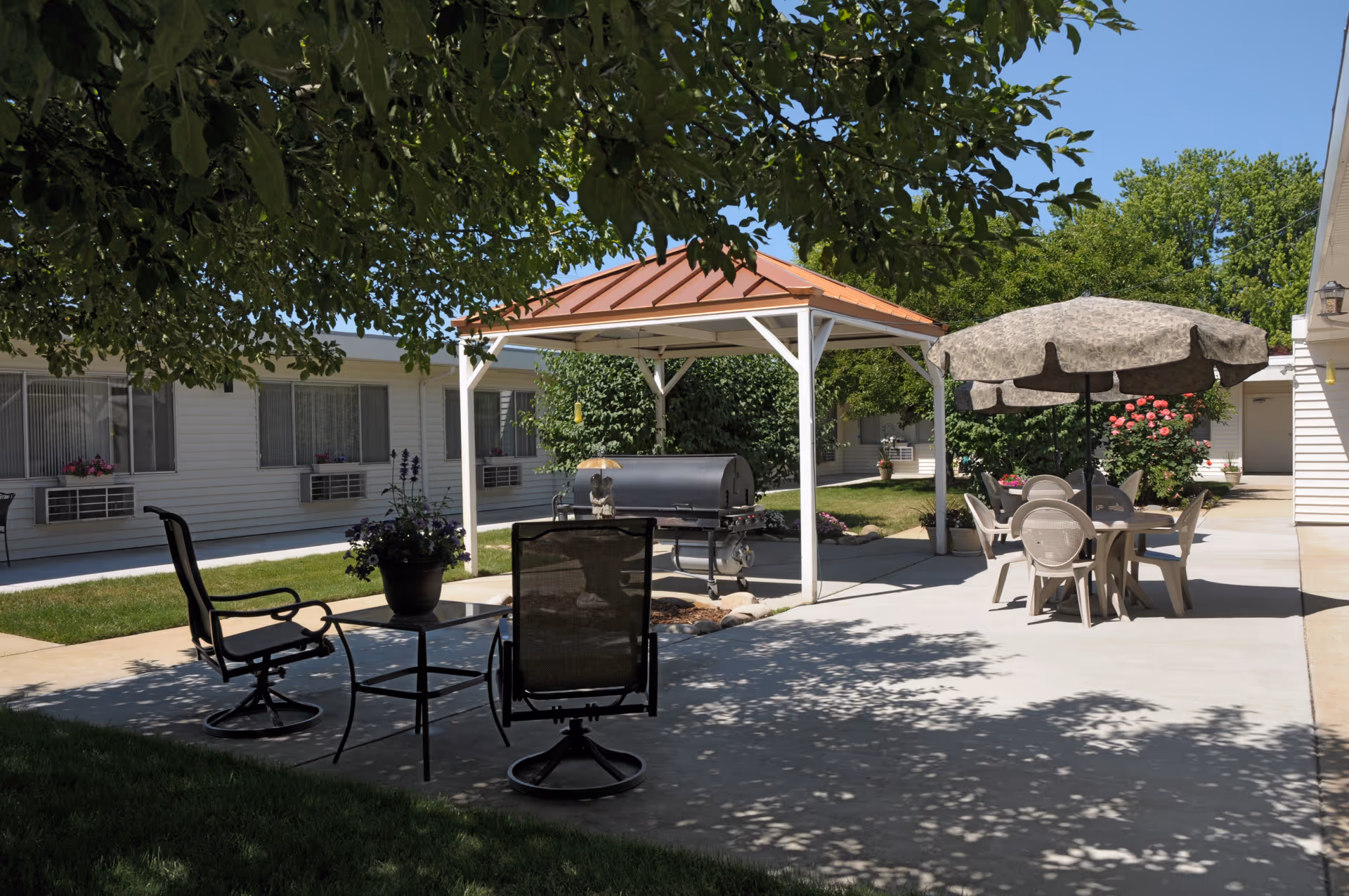 Outdoor patio area at a senior living facility with a covered grill, several chairs and tables, an umbrella providing shade, and surrounding greenery including trees and flowering bushes.