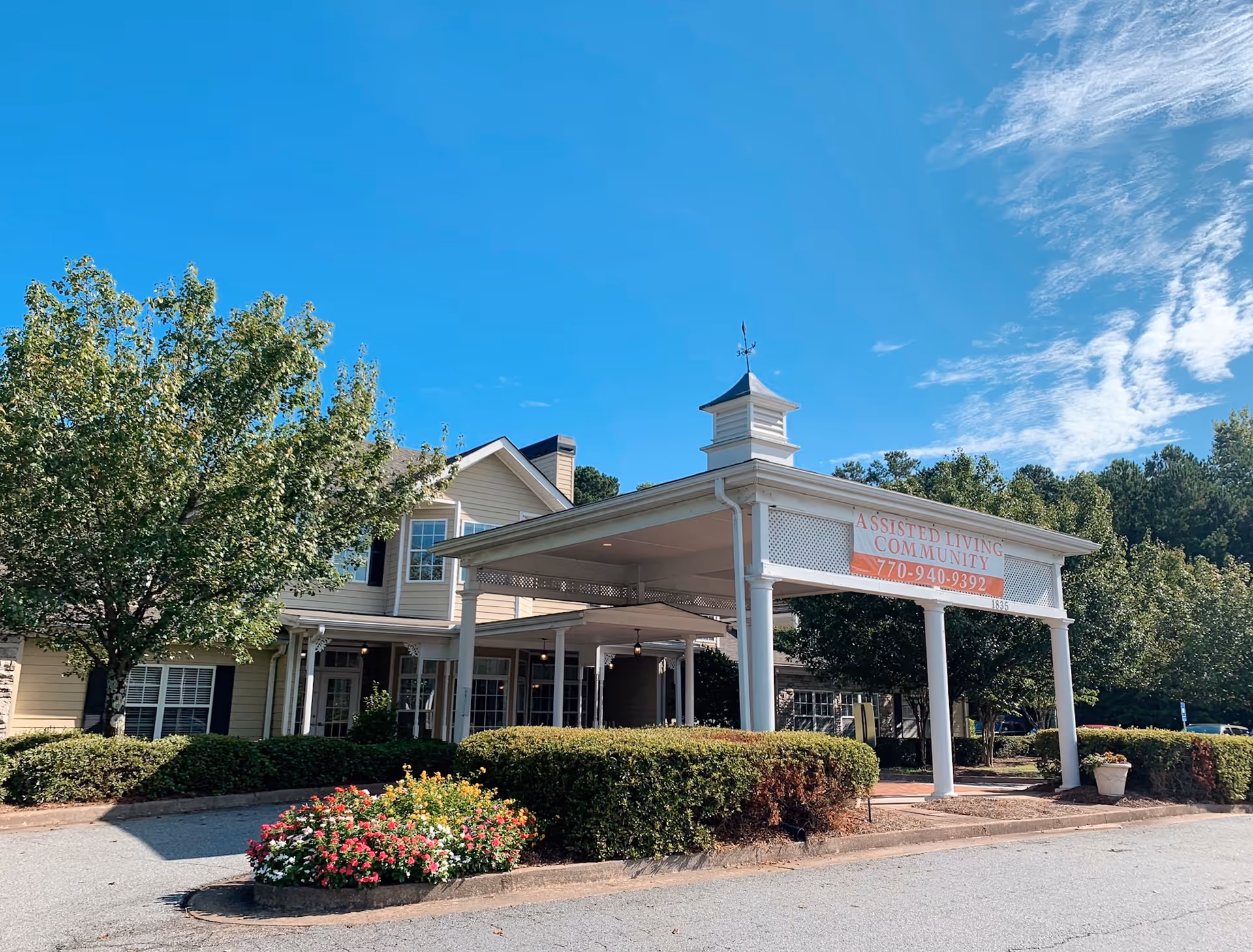 Exterior view of a senior living facility building with a covered entrance supported by white columns. There are bushes and a flower bed with colorful flowers in front of the building. The sky is clear and blue with some clouds.