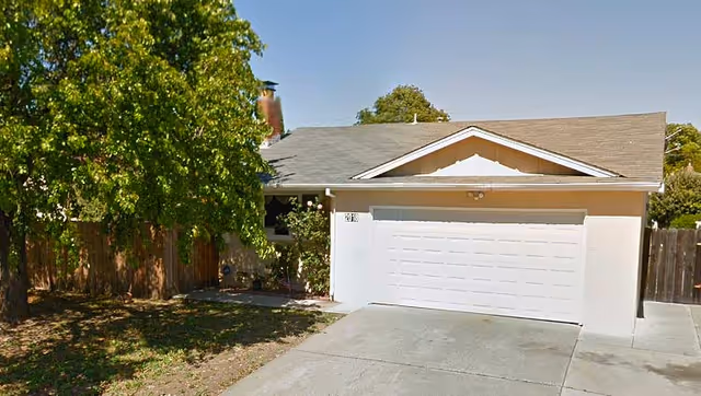Front exterior view of a single-story residential house with a two-car garage, a tree on the left side, and a driveway leading up to the garage.