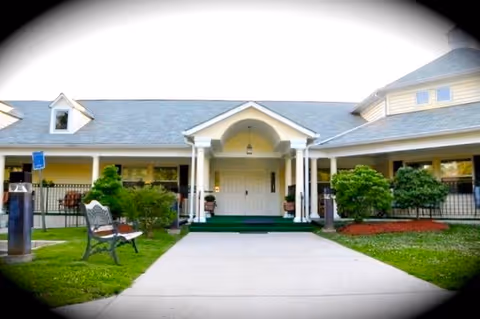 Front entrance of a single-story elder care facility with a covered porch supported by white columns, a concrete walkway leading to double doors, green grass, bushes, and a bench on the left side.