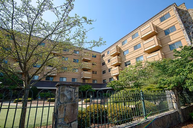 Exterior view of a multi-story senior living facility with balconies, trees, a landscaped courtyard and a metal fence.