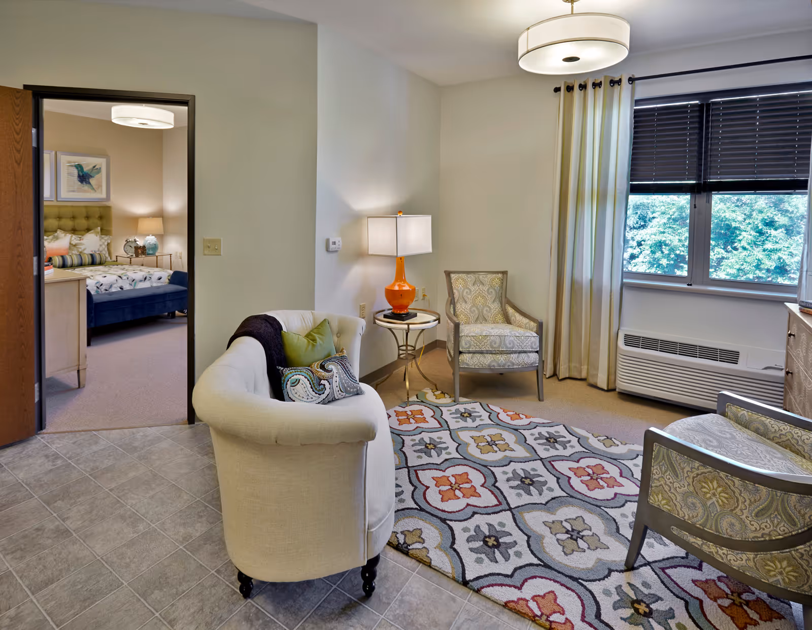 A cozy living room area with a beige curved sofa adorned with pillows, two patterned armchairs, a colorful patterned rug, a side table with an orange lamp, and a large window with curtains and blinds. Through an open doorway, a bedroom with a bed, nightstand, and wall art is visible.