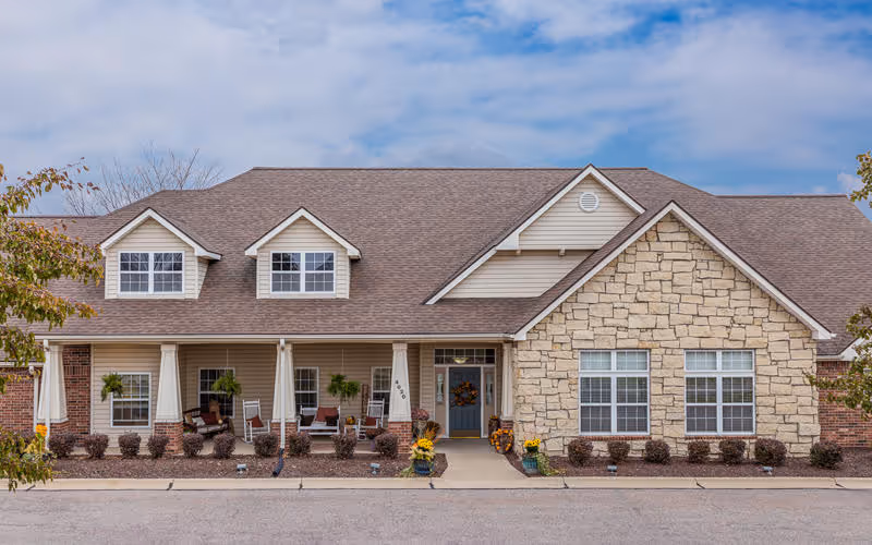 Front exterior view of a single-story building with a combination of stone and siding facade, multiple windows, a covered porch with seating, and decorative plants and flowers near the entrance under a partly cloudy sky.