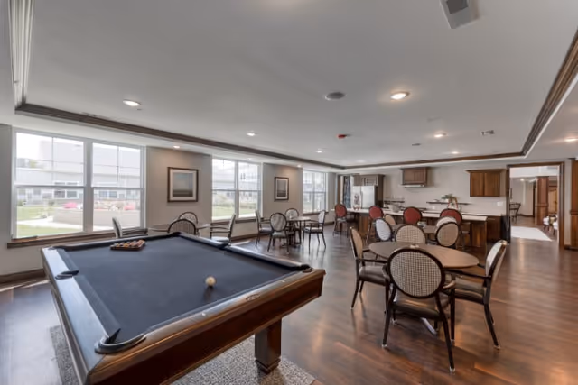 A spacious common area with a pool table in the foreground and several round tables with chairs arranged throughout the room. Large windows let in natural light and provide a view of an outdoor garden area. The back of the room features a kitchen area with wooden cabinets and a refrigerator.