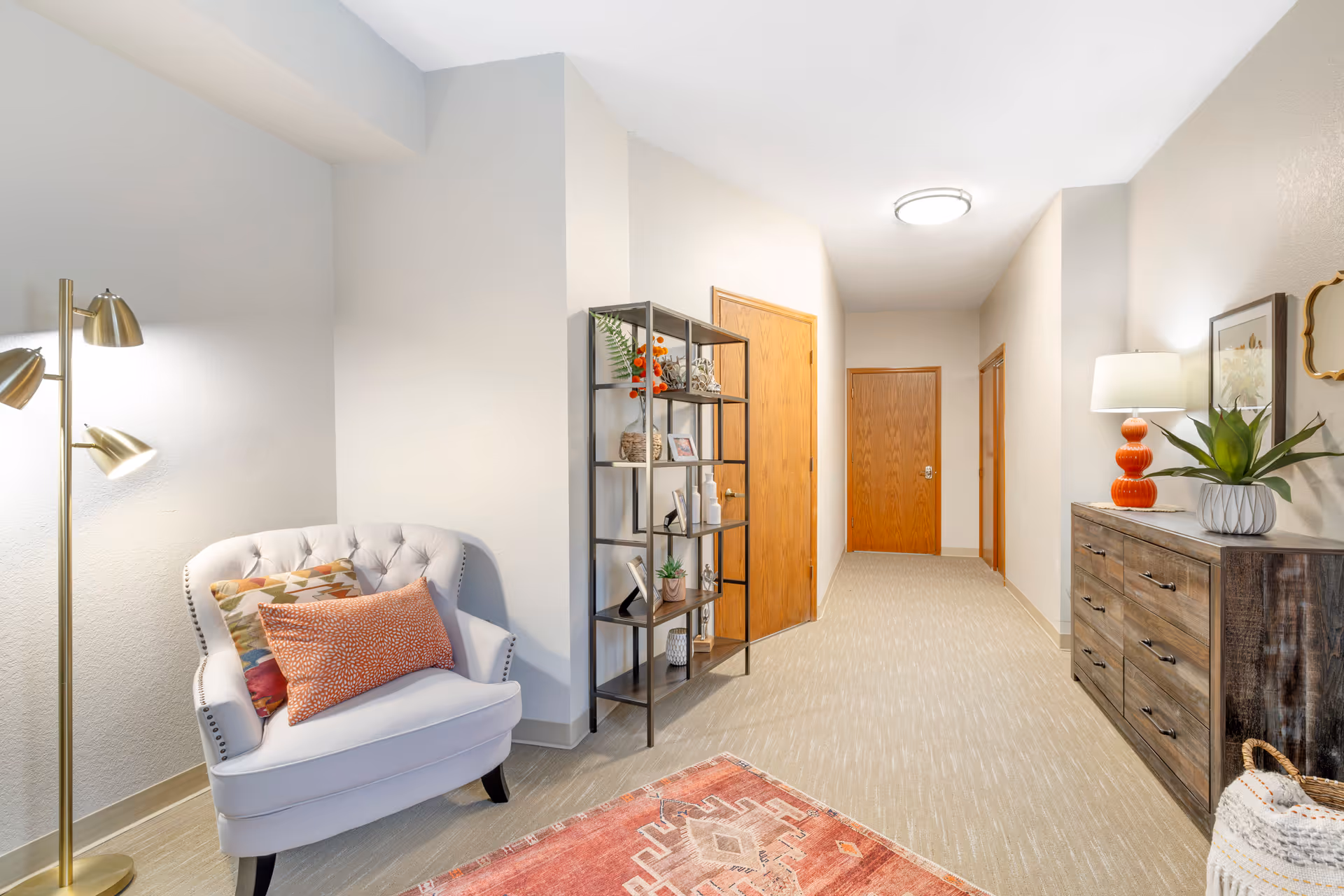 A well-lit hallway in an assisted living facility with beige walls and carpeted floor. On the left side, there is a white upholstered armchair with two decorative pillows and a gold floor lamp. Next to the chair is a black metal shelving unit with various decorative items. On the right side, there is a wooden dresser with a red lamp, a potted plant, and framed artwork above it. The hallway has three wooden doors and a ceiling light fixture.