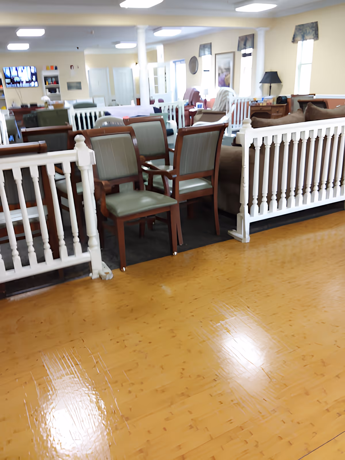Interior view of a senior living facility common area with wooden flooring, several chairs with green cushions, white wooden railings, sofas, and a television mounted on the wall in the background.