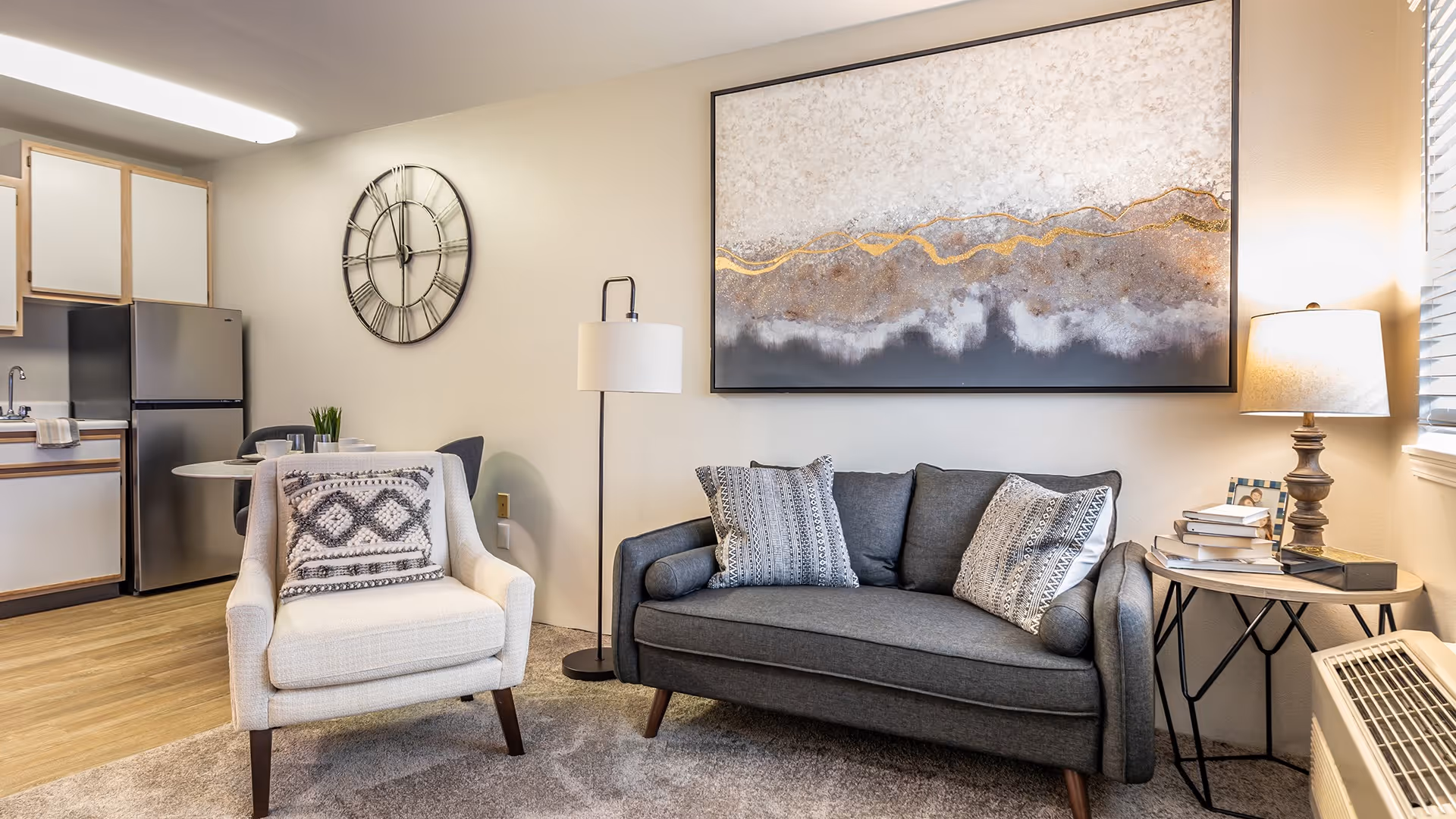 A cozy living room area with a gray sofa adorned with patterned pillows, a white armchair with a decorative pillow, a floor lamp, and a side table with a lamp and books. Behind the seating area is a large abstract painting. To the left, there is a small kitchen area with a refrigerator, cabinets, and a round dining table with chairs. The room has wooden flooring and a large wall clock.
