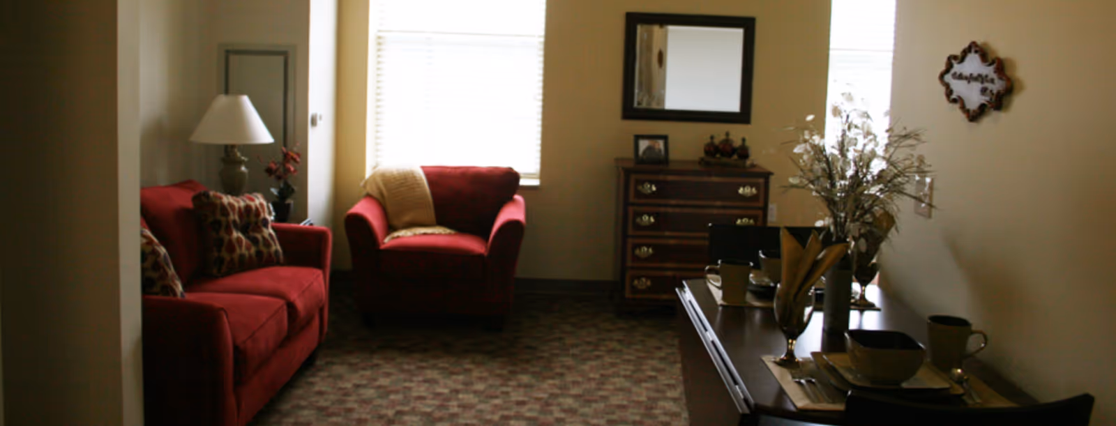 A cozy living room area with a red sofa and matching armchair, both adorned with patterned cushions. A beige throw blanket is draped over the armchair. There is a table lamp on a side table next to the sofa, and a wooden dresser with a mirror above it is against the far wall. A small dining table with place settings and a vase with flowers is visible in the foreground. The room has carpeted flooring and two windows with blinds letting in natural light.