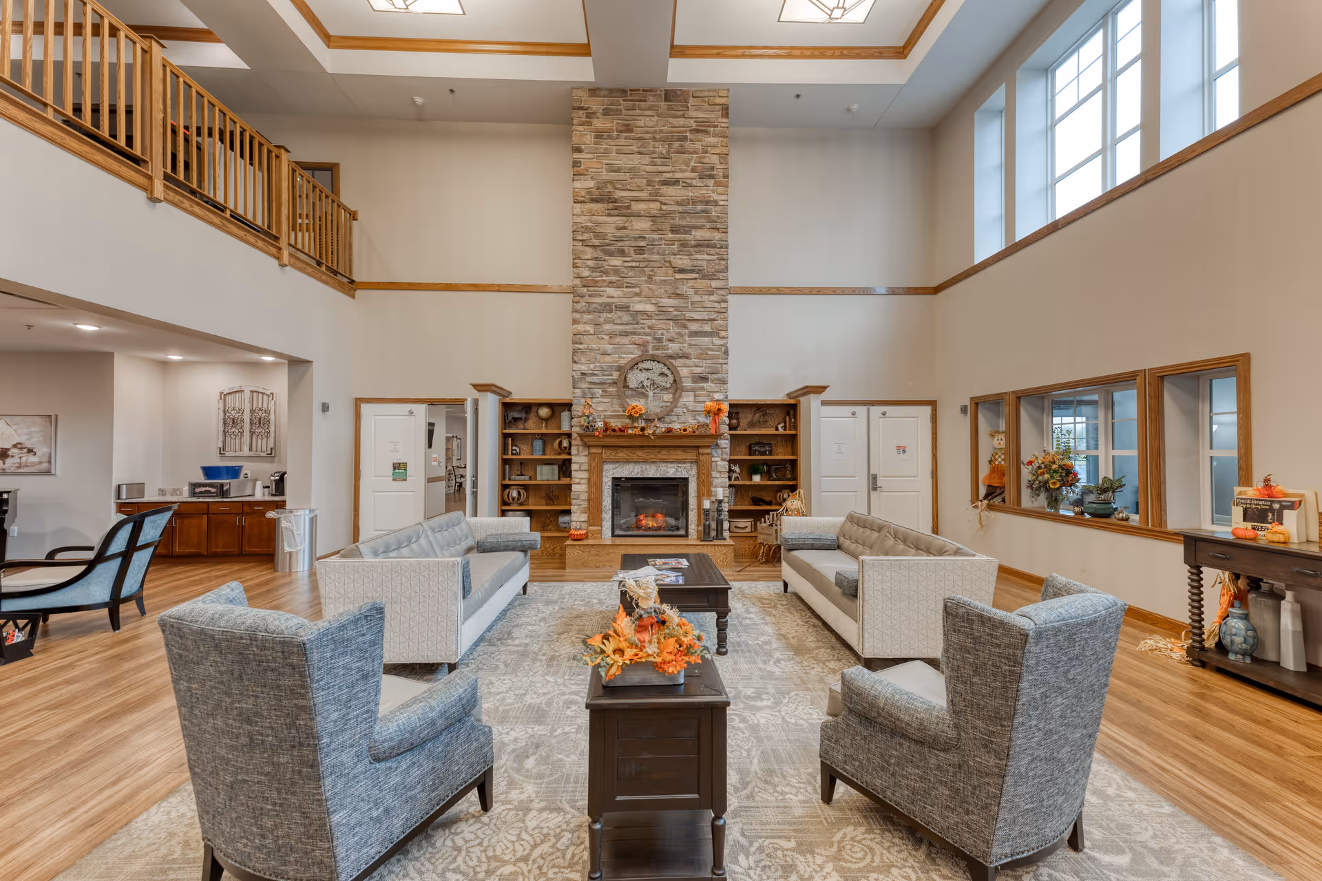 A spacious senior living facility common area with high ceilings and large windows. The room features a stone fireplace centered on the far wall, flanked by wooden bookshelves. There are two light gray sofas and two blue armchairs arranged around a dark wooden coffee table on a patterned area rug. The room has wood flooring and light-colored walls with wooden trim. Decorative fall-themed accents are placed on the tables and fireplace mantel.
