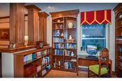 Interior view of a cozy room featuring wooden bookshelves filled with books and decorative items, a wooden desk with a computer, a wooden chair with a green cushion, and a window with a red and yellow valance.