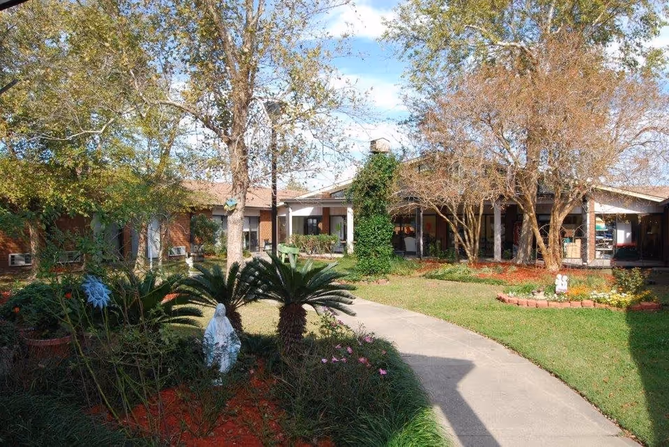 Curved walkway leading through a landscaped courtyard with palm-like plants, trees, and a single-story building entrance.