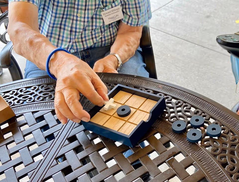 An elderly person wearing a blue and white checkered shirt and a blue wristband is playing a game of tic-tac-toe on a decorative metal outdoor table. The person is holding a white cross game piece above the wooden tic-tac-toe board, which also has black circular pieces on it and scattered on the table. The setting appears to be outdoors on a concrete surface.