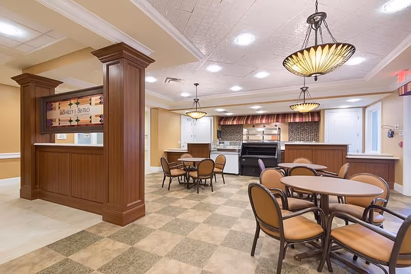 Interior view of a dining area in a senior living facility with round tables and chairs arranged on a checkered tile floor. The room features warm beige walls, decorative hanging light fixtures, and a serving counter area with a sign that reads 'Bailey's Bistro'.