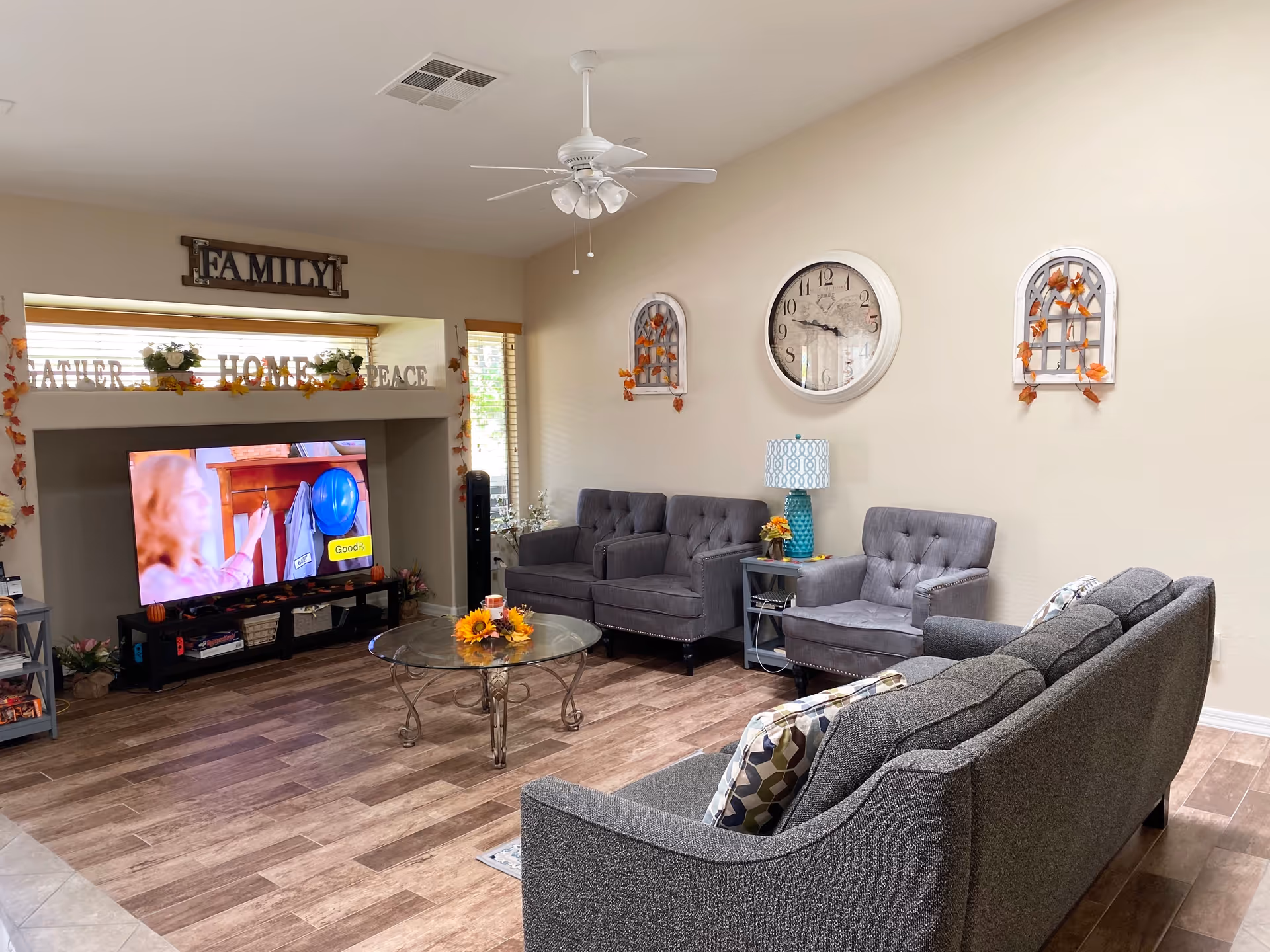 A cozy living room with a gray couch and two gray armchairs arranged around a glass coffee table with a floral centerpiece. A large wall clock and decorative wall hangings with autumn leaves are on the beige wall. A ceiling fan with lights is mounted on the ceiling. A TV is placed in a recessed area with decorative items on the mantle above it, including the words FAMILY, FATHER, HOME, and PEACE. The room has wood-look flooring and window blinds letting in natural light.