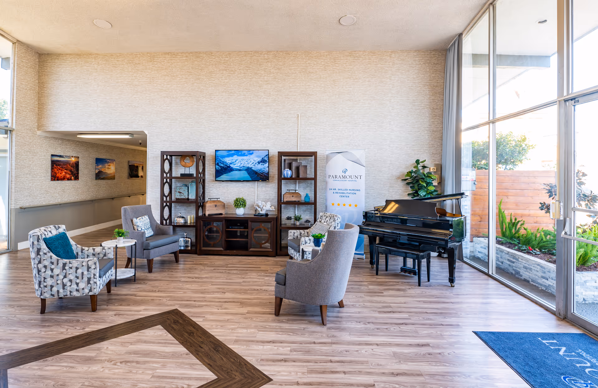 Bright lobby of Paramount Convalescent Hospital showing a seating area with chairs, a grand piano, shelving, a TV, and large floor-to-ceiling windows.
