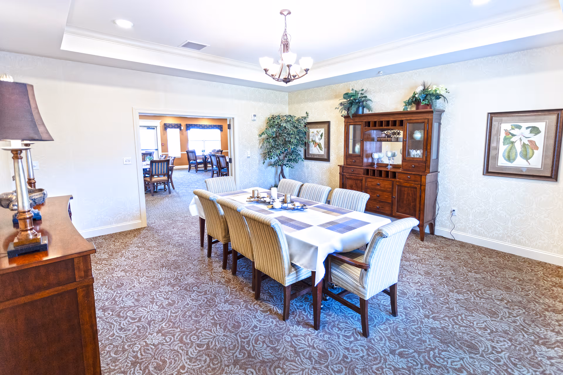 A well-lit dining room with a long table covered with a white tablecloth and placemats, surrounded by eight upholstered chairs. The room features patterned carpet, a wooden sideboard with a lamp on the left, a wooden china cabinet with decorative items on the right, framed botanical artwork on the walls, and a potted plant in the corner. Through an open doorway, another dining area with tables and chairs is visible.