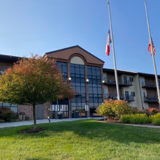 Exterior view of a multi-story senior living facility with large windows and balconies. There are two flagpoles with flags at half-mast in front of the building, a well-maintained lawn, and a tree with autumn-colored leaves.