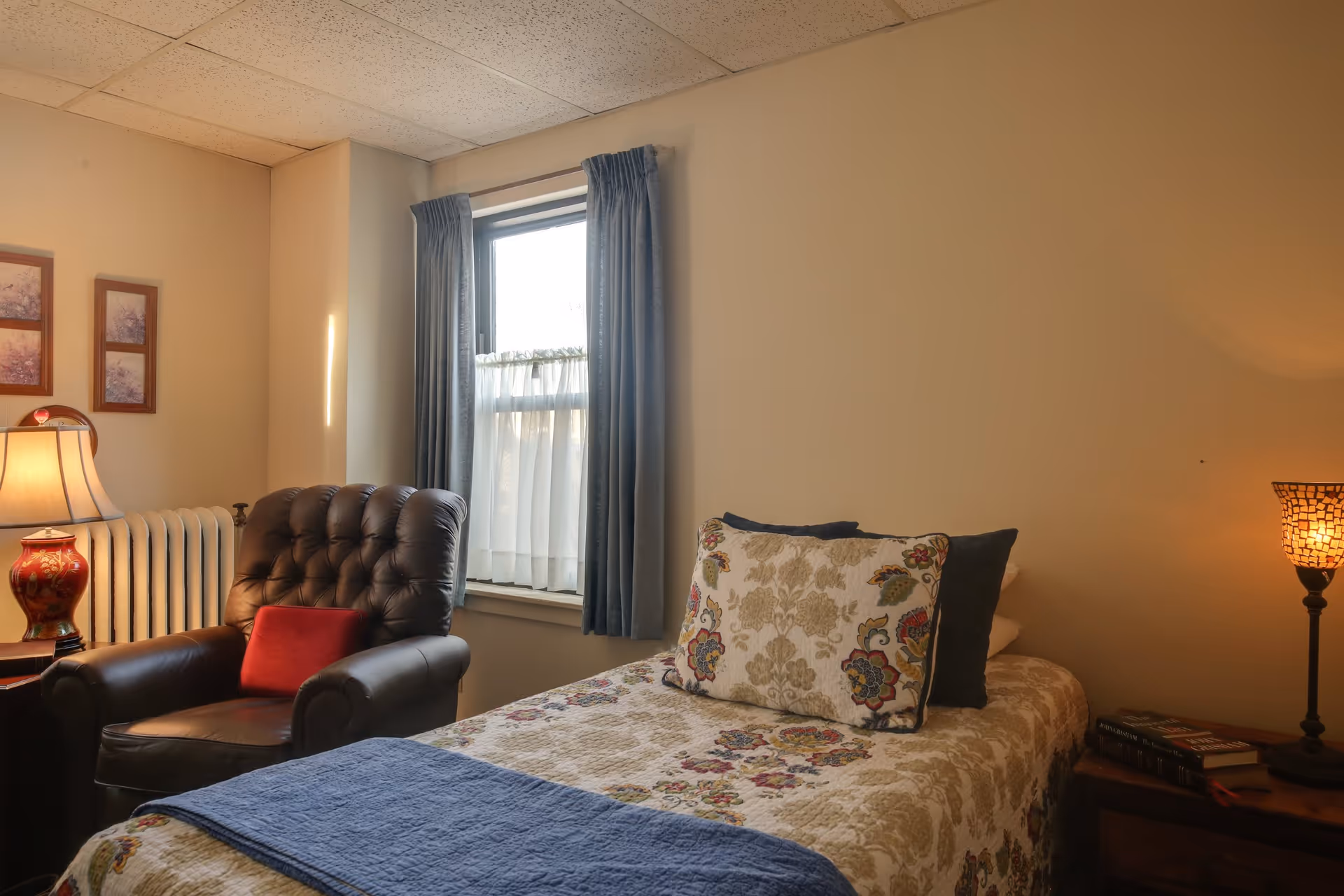 A cozy bedroom with a single bed covered in a floral patterned quilt and pillows. Next to the bed is a wooden nightstand with a lit lamp and a few books. There is a dark brown leather armchair with a red cushion beside a window with blue curtains. The walls are beige and decorated with two framed pictures.