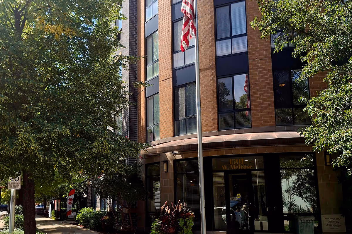 Front of a brick multi-story senior living building with large windows, an entry door, a flagpole, and trees along the sidewalk.